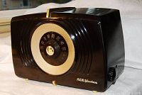 Front angled view of brown Bakelite table radio with circular dial, gold-toned speaker grille and feet, RCA Victor logo on front.