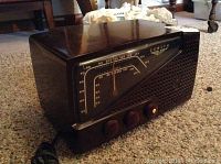 Front view of the Zenith radio showing its dark brown plastic casing, tuning dial with AM/FM frequencies, and three dials. Visible minor crack on the front cover's left side and missing brand logo.