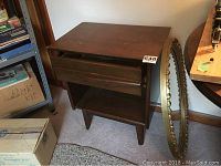 Vintage Gibbard end table in solid wood, one drawer and lower shelf. Shows wear and veneer chipping on left front corner. Oval mirror with gold painted plastic frame leaning against wall beside table.