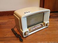 Front angled view showing ivory plastic casing, speaker grill, tuning dial with frequency numbers, and knobs with power cord.