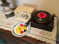 Photo shows RCA Victor retro radio with built-in clock and RCA Victor turntable with 45 RPM record on the platter, plus a plate with various plastic 45 adapter pieces on a lace cloth-covered surface.