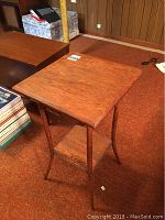 Vintage oak occasional table shown from the front, highlighting its square top and curved legs over lower shelf, positioned on carpeted floor with other furniture in background.