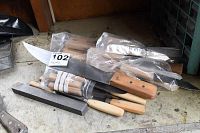Various handmade knives with wooden handles and metal blades, some bagged, arranged on a surface with a sharpening stone