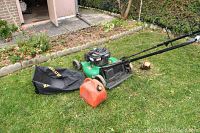 Green Briggs and Stratton lawn mower with black grass catcher and orange fuel can placed on grass near garden bed and pathway.