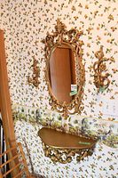 Full view of the gold ornate oval mirror flanked by two matching gold wall sconces and the wall-mounted half-moon table below it, against floral wallpaper.