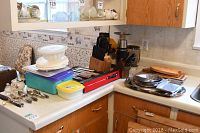 View of kitchen corner countertop showing stacked plastic storage containers with lids, stainless steel cutlery set, wooden knife block with knives, metal trays, and a ceramic sugar bowl.