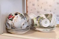 Photo shows approximately 10 assorted teacups and matching saucers arranged in two stacks. Cups display floral designs with gold trim, showcasing brands like Aynsley, Paragon, and Queen Anne.