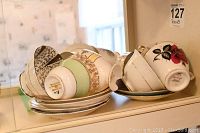 Eleven various patterned tea cups and approximately nine matching saucers stacked together showing different colors and designs including a cup with a broken base.