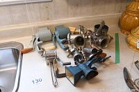 Overview of eight vintage metal kitchen tools and grinders placed on countertop near sink, showing variety of shapes, sizes, colors, and materials including metal and wood handles.