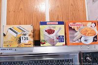Photo showing three boxed kitchen appliances: Atlas pasta maker, Leifheit cherry stoner, Soehnle kitchen scale lined up atop an oven.