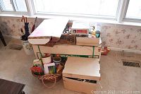Boxes containing assorted Christmas ornaments and an artificial tree box stacked near a window with visible floral wallpaper.
