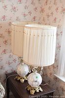 Pair of decorative table lamps on wooden surface, showing overall height, spherical floral glass bases with gold-tone metal details and cream pleated shades