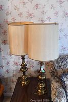 Pair of brass table lamps with cream-colored fabric shades on wooden side table next to floral upholstery couch and wallpaper.