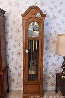 Full view of the tall grandmother clock against floral wallpaper, showing wood finish and glass door revealing the pendulum.