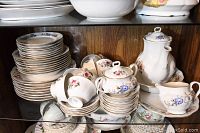 Stack of plates, tea cups, saucers, sugar bowl, creamer, and teapot with floral patterns and gold trim in a wooden cabinet.