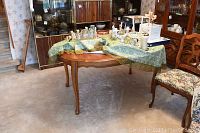Oval wooden dining table with a green patterned cloth with items on top, showing carved legs and wood finish.