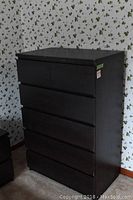Photo of dark wood and veneer dresser with six drawers against a floral wallpaper background.