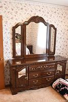 Front view of large wood framed dresser with carvings and attached triple mirror showing the floral wallpaper background