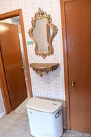 View of ornate gold plastic framed wall mirror, matching wall shelf, and white wicker hamper beneath, all arranged in a hallway corner.