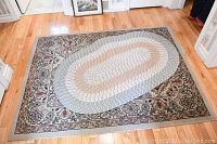 Photo showing the two rugs stacked, with the smaller oval braided rug on top of the larger rectangular floral border rug on a hardwood floor.