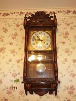 Full front view of the ornate carved wooden wall clock mounted on floral wallpaper background, showing overall clock design and pendulum through the glass door.