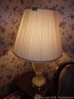 One of the crystal table lamps illuminated on a dark wood table with floral wallpaper background, showing pleated beige fabric shade and crystal base with round metal bottom.