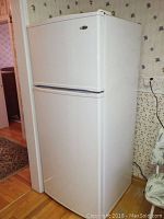 Full side and front view of white Amana top freezer refrigerator placed against wall indoors on hardwood floor.