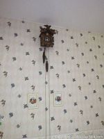 Photo of the Lindner Handarbeit Germany cuckoo clock mounted on floral wallpapered wall along with two small framed floral prints below it.
