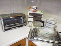 Photo showing Hamilton Beach toaster oven on countertop with metal baking pans and rack stacked beside it, also showing white Pyrex casserole dishes with lids and Mr. Coffee machine partially visible.