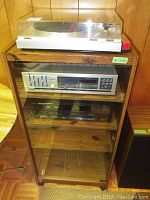 Full view of laminate wood cabinet with glass front showing turntable on top, cassette deck and synthesizer inside shelves.