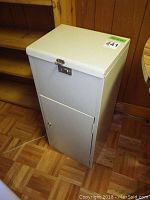 Photo of small white metal cabinet standing on wooden floor, showing hinged top and bottom door