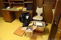 Wide shot showing black photo enlarger, metal developing tank, boxes with photo paper and accessories, wooden base board, chair, desks, shelves and broom.
