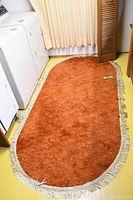 Orange oval rug about 6 feet long with white fringe trim displayed in laundry room against wall and washer/dryer.