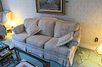 Beige fabric couch with floral patterns and two matching cushions, placed against wall under a painting and near a lamp and chair.