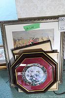 Photo showing three framed art prints stacked behind an octagonal framed decorative plate with floral and hummingbird pattern, all on floor against wallpapered wall.