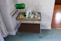 Heated serving cart made of metal and wood holding an electric clothes iron, a green glass lamp, and a power cord on a carpeted floor against wallpapered wall.