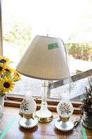 Three vintage lamps on a wooden surface near a window, two with floral milk glass shades and one larger lamp with a fabric pleated shade.