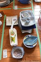 Top view showing multiple stacked white ceramic platters, blue fish-shaped plates, square purple-yellow floral bowl, hand-shaped utensil, and corn cob shaped butter dish.