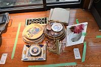 Photo showing multiple decorative plates stacked, a framed floral print, a rectangular tray, and a triangular metal candle holder all placed on hardwood floor.