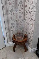 Photo showing clear glass vase seated on wooden hexagonal stand against floral-patterned wallpaper.