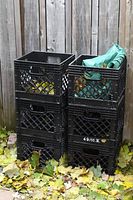 Six black plastic milk crates stacked in two columns outdoors near wooden fence, green tarp folded in top right crate, dry leaves on ground.