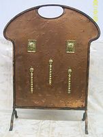 Front view of the copper fire screen showing embossed patterns, three brass decorative nails, two brass sunburst studs near the top, and handle cutout at top center.