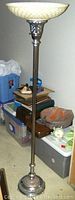 Full view of the tall chrome floor lamp standing near stored items, displaying the overall height and slender pole with the wide glass shade at the top.