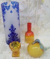 Four art glass pieces including tall frosted blue cameo vase, amberina vase, smaller amber glass stand, and Murano glass bird figurine on a white patterned cloth