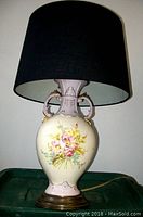 Porcelain lamp with floral design and black lampshade shown from the front, placed on a green surface.