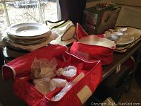Image showing boxed and wrapped holiday dishes, multiple white dish pieces stacked with protective fabric and bubble wrap in red storage cases.