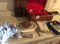 Flatware sets displayed on a countertop including a wooden box with Tudor Plate Oneida silverware, bundled stainless steel flatware wrapped in plastic, and decorative hexagonal plates.