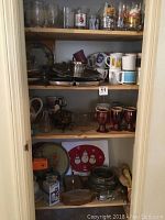 Shelving unit holding several kitchen and serving pieces including brown Hull mugs, collectible glasses, stainless steel utensils, and decorative plates