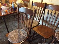 Four brown stained wooden dining chairs with spindle backs and turned legs. Two chairs have round braided cushions tied on the seats. Chairs show slight surface imperfections and nicks.