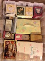 Multiple Precious Moments figurines in boxes seen inside a plastic bin, including a box with pastel patterns resembling a tissue box.